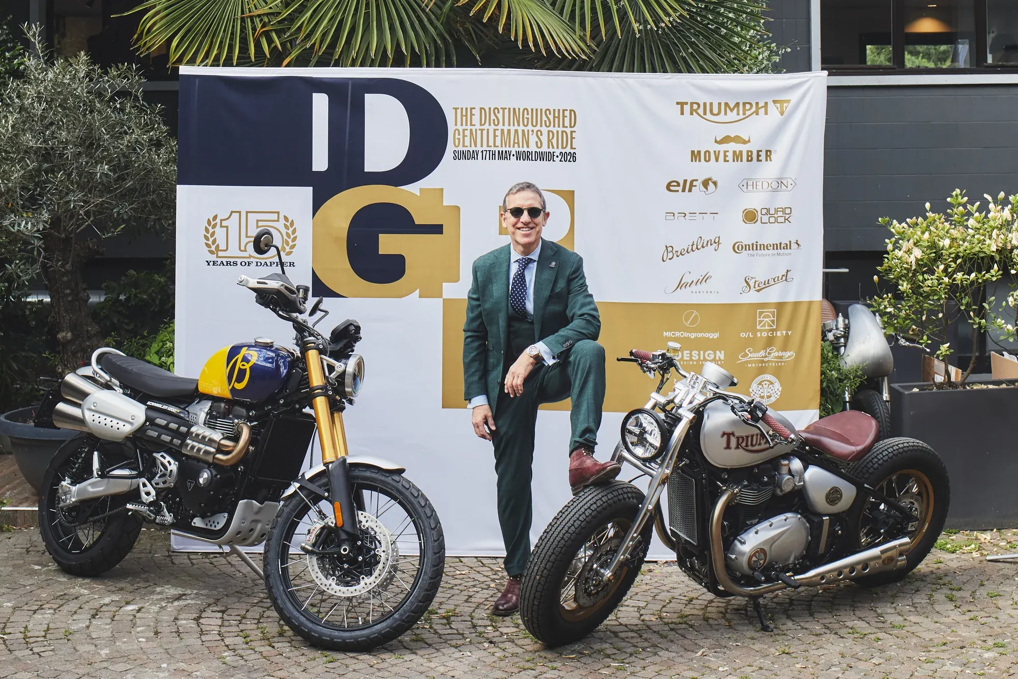 Matteo Adreani al punto di ritrovo del Distinguished Gentleman's Ride Milano 2026: completo verde scuro tre pezzi, occhiali da sole, in piedi fra due moto Triumph davanti al backwall DGR con il logo dei quindici anni di customizer e i loghi degli sponsor