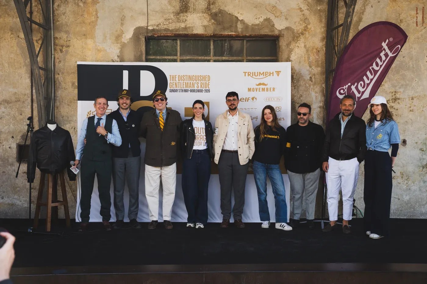 Sul palco dello Spirit de Milan al termine del Distinguished Gentleman's Ride Milano 2026: il team allineato in giacca, cravatta e gilet, sfondo industriale con il muro patinato, backwall DGR e bandiera Stewart's, una giacca in pelle esposta sul manichino a sinistra