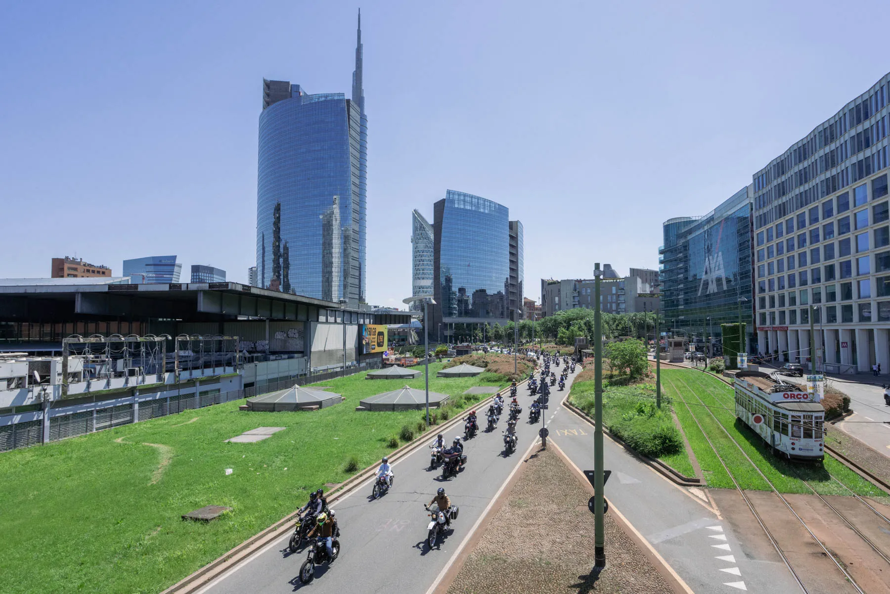 Una colonna di moto classiche con motociclisti vestiti in giacca e cravatta in partenza per una via di Milano, luce di mattina